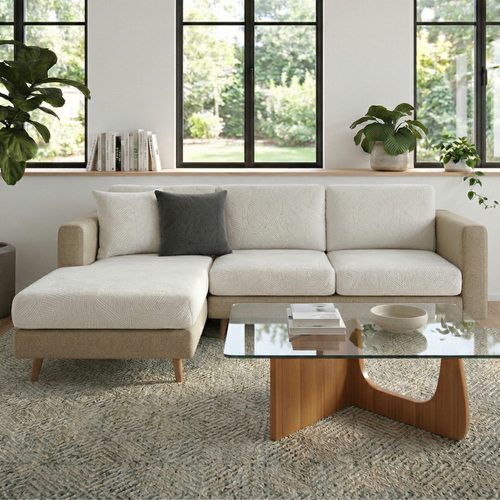 Beige sectional sofa in a living room with a textured jacquard leaf white sofa cover, and glass coffee table and large windows.