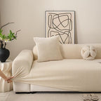 Beige sofa with a person stretching a matching beige fabric over it, in a room with abstract art and plants.