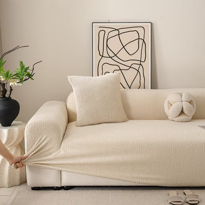 Beige sofa with a person stretching a matching beige fabric over it, in a room with abstract art and plants.